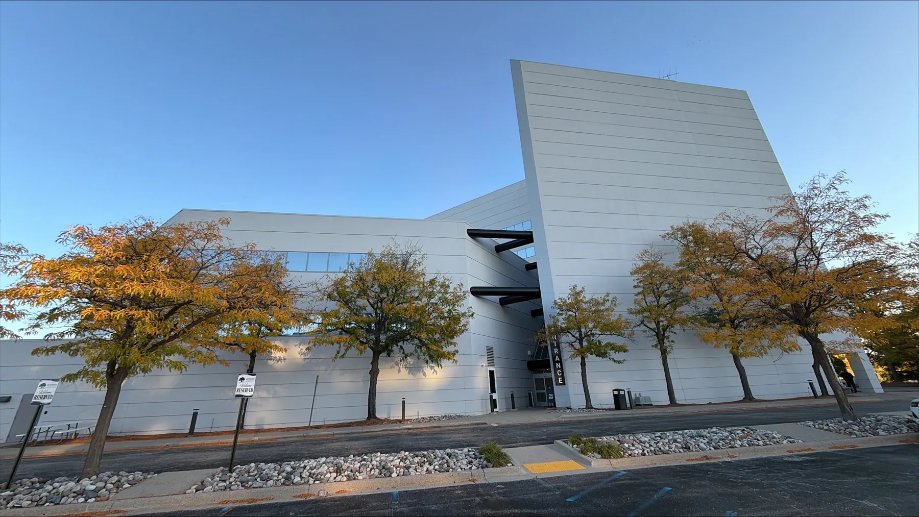 The white, angular office building at 801 W. Big Beaver Road in Troy, with a tall vertical Entrance sign beside the glass front doors, a row of autumn trees along the walkway and two reserved parking signs at the curb in the foreground