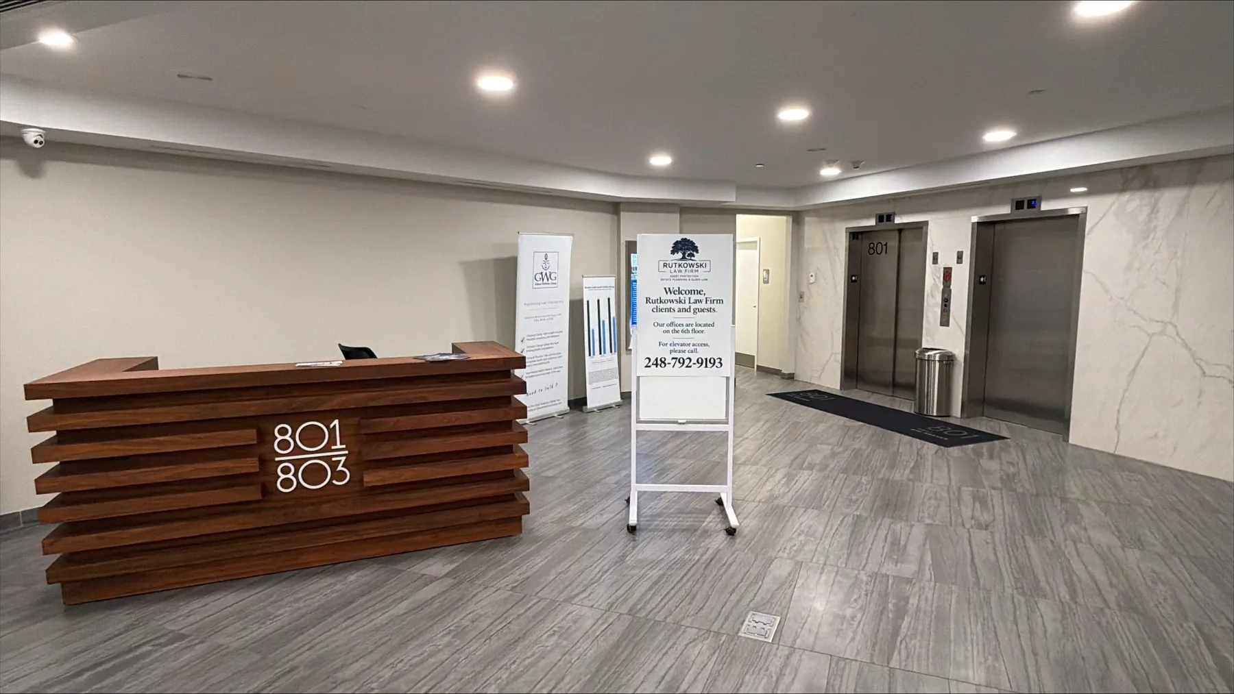 The ground-floor lobby at 801 W. Big Beaver Road, with a wood-slat front desk marked 801/803 on the left, a Rutkowski Law Firm welcome sign on an easel in the center listing the elevator phone number 248-792-9193, and two stainless steel elevators on the right