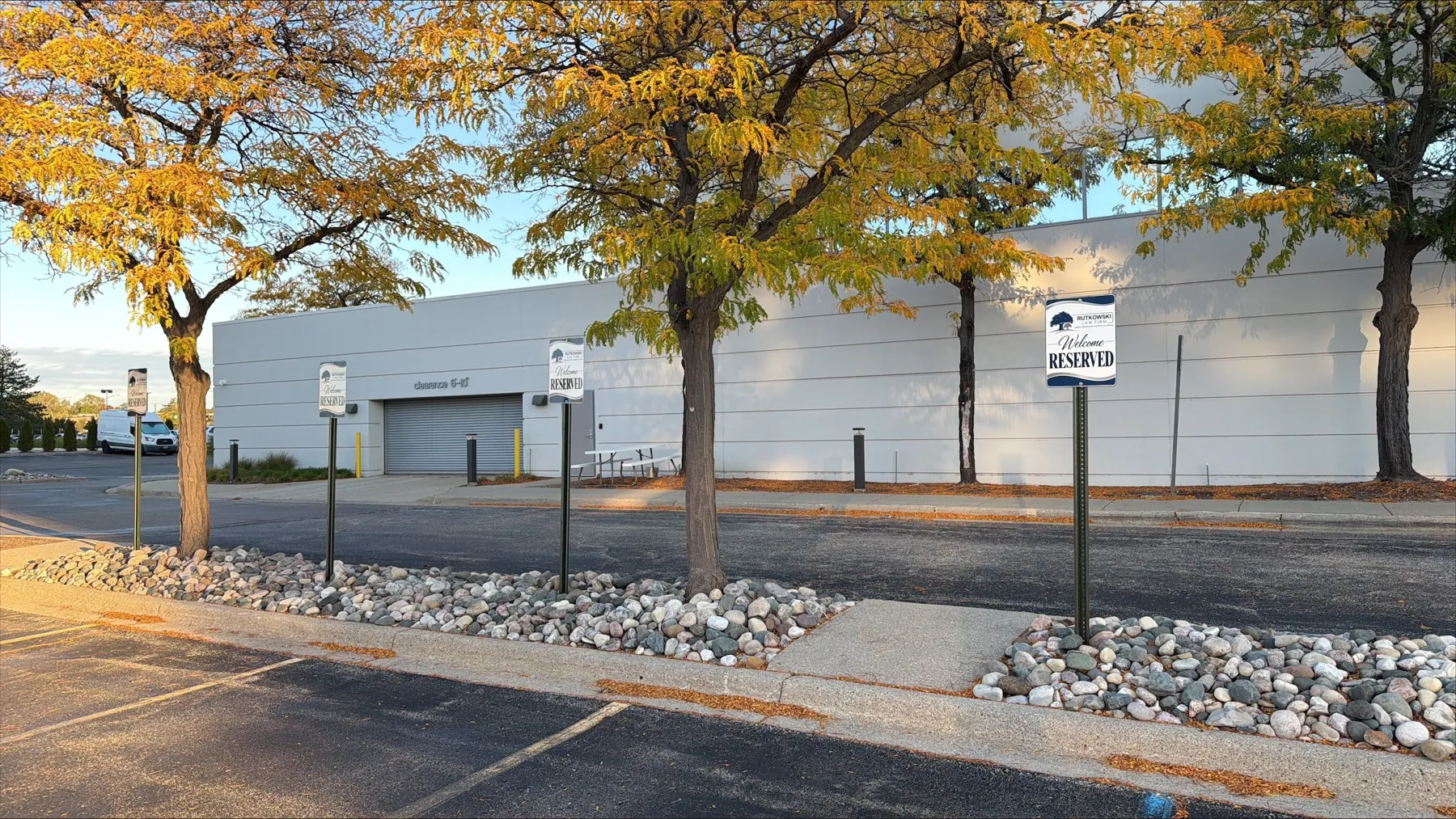 Three blue and white Rutkowski Law Firm "Welcome, Reserved" parking signs on posts along a rock-lined curb island outside 801 W. Big Beaver Road in Troy, with yellow autumn trees and the building's white side wall behind them