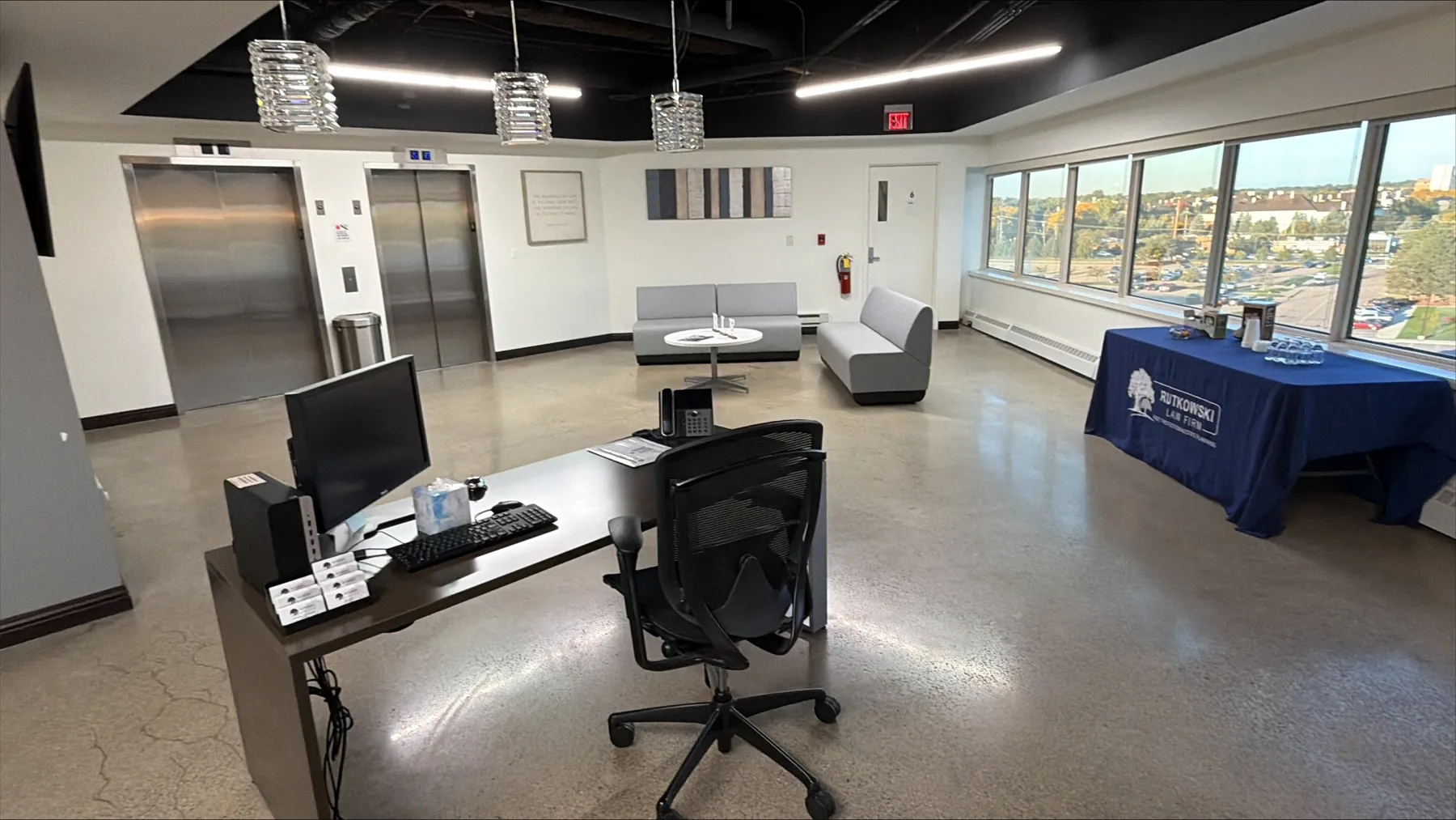 The sixth-floor elevator lobby at Rutkowski Law Firm's Troy office, with two stainless steel elevators along the back wall, a reception desk with a computer and phone in the foreground, gray lounge seating, and a navy Rutkowski Law Firm table beneath a wall of windows overlooking Troy