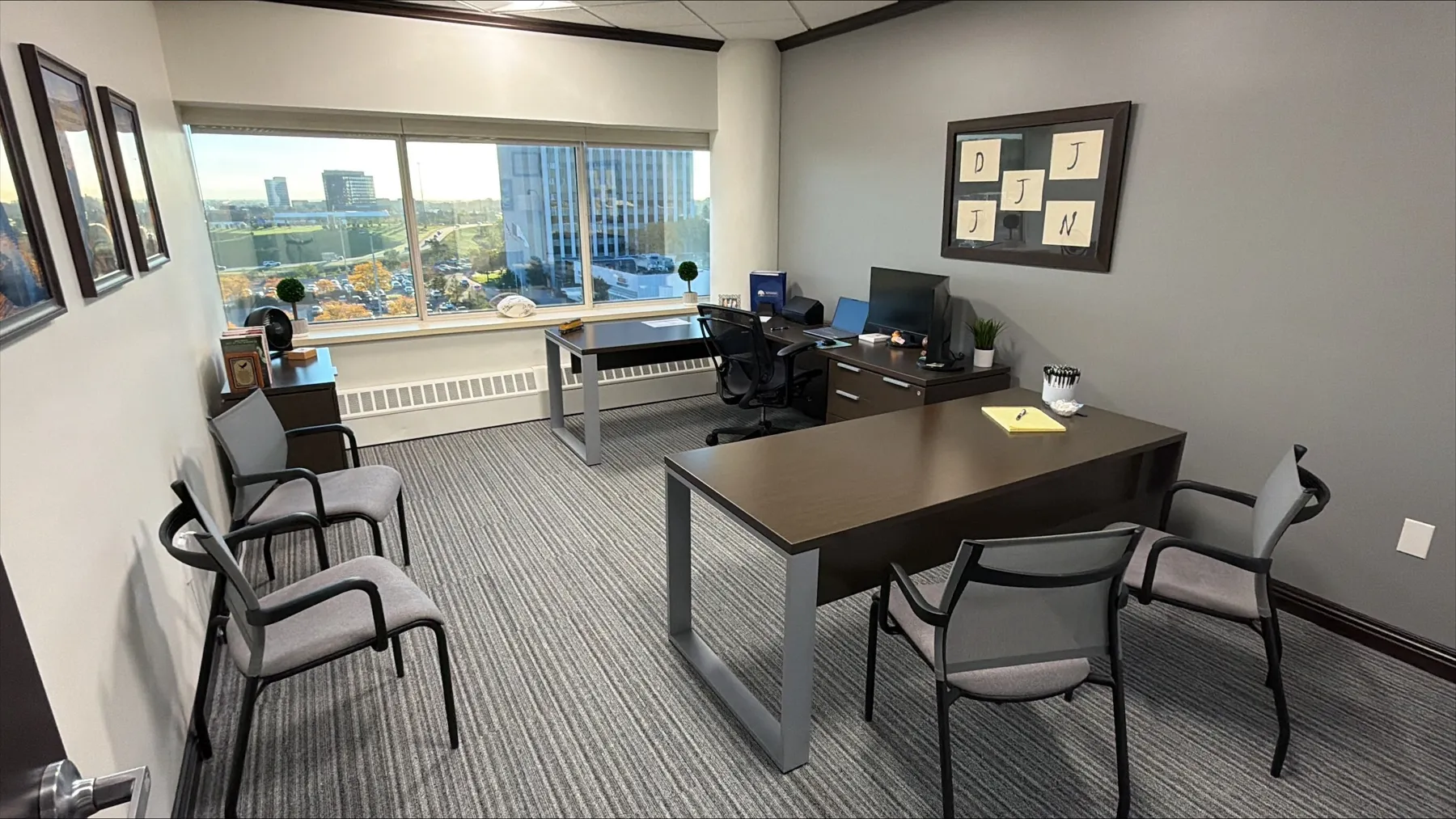 A Vision Consultation office at Rutkowski Law Firm in Troy, with a dark wood desk facing four gray mesh guest chairs, framed photos on the left wall, a corkboard of sticky notes on the right wall, and a wide window looking out over Big Beaver Road, its parking lots and the neighboring office towers